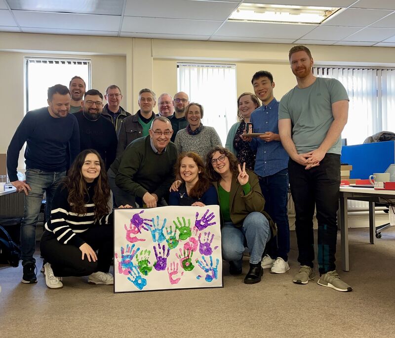 A photo shows a group of smiling people behind a handprint poster made in the colours of rare disease day